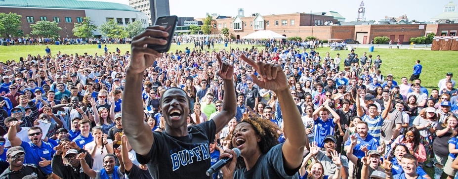 Large group of students outside on campus, many wearing UB blue, and two n front taking a selfie together. 
