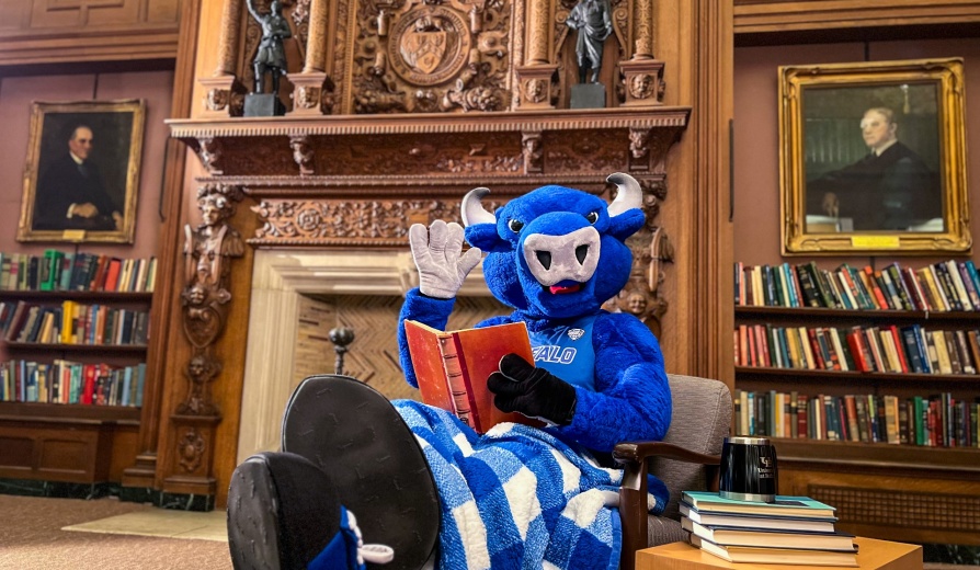 Victor E. Bull, the UB mascot, reads a book near the fireplace in Abbott Library in December 2022. Photographer: Douglas Levere. 