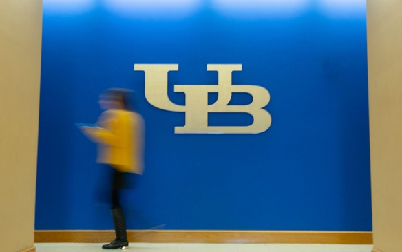 UB logo on a wall with a student walking in front of it. 