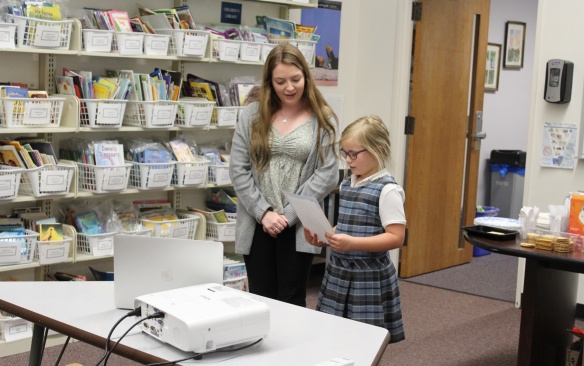 A student reading in front of the class with her tutor. 