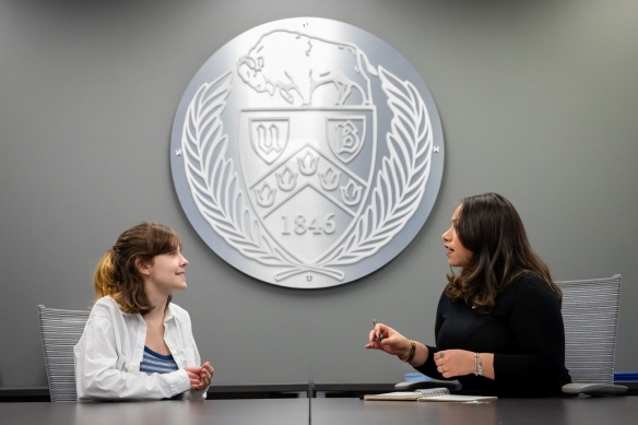 Two faculty meeting in front of the UB seal on a wall. 