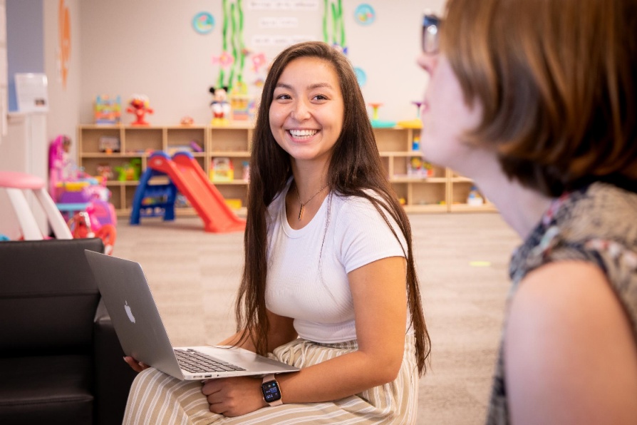 Student sits in childrens' classroom with a laptop in lap, smiling at teaching instructor. 