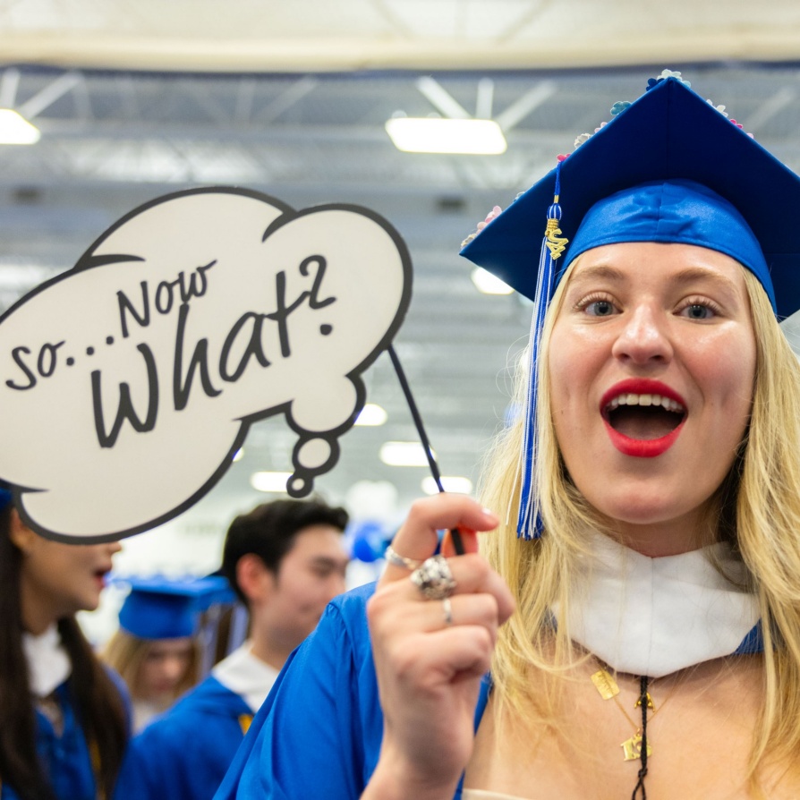 Graduate at commencent in cap and gown holding up a sign that reads So...No What? 