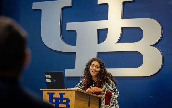 Speaker at a podium in front of a large UB logo. 