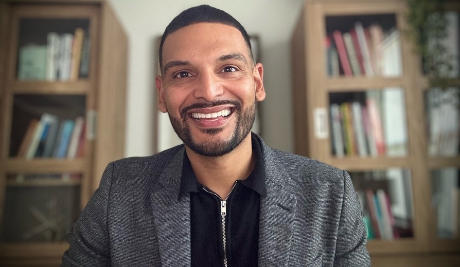 Stephen Santa-Ramirez smiles brightly while wearing gray blazer and black shirt. Bookshelves are in the background. 