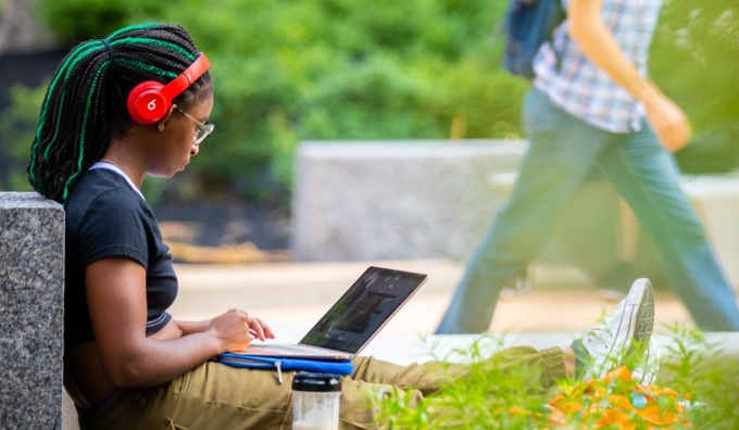 Student using laptop outside on campus, wearing red headphones, with green foliage nearby. 