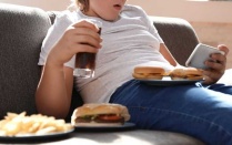 Child eating burgers and drinking soda on a couch while watching a phone. 
