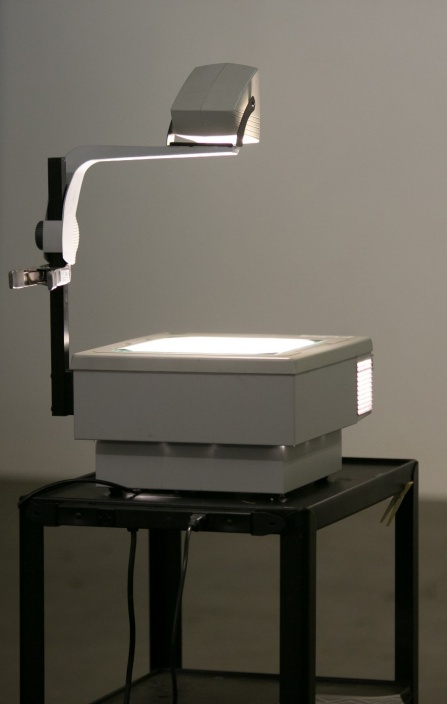 A vintage overhead projector sitting on a roller cart lighting a wall ready to show overhead projection transparencies. 