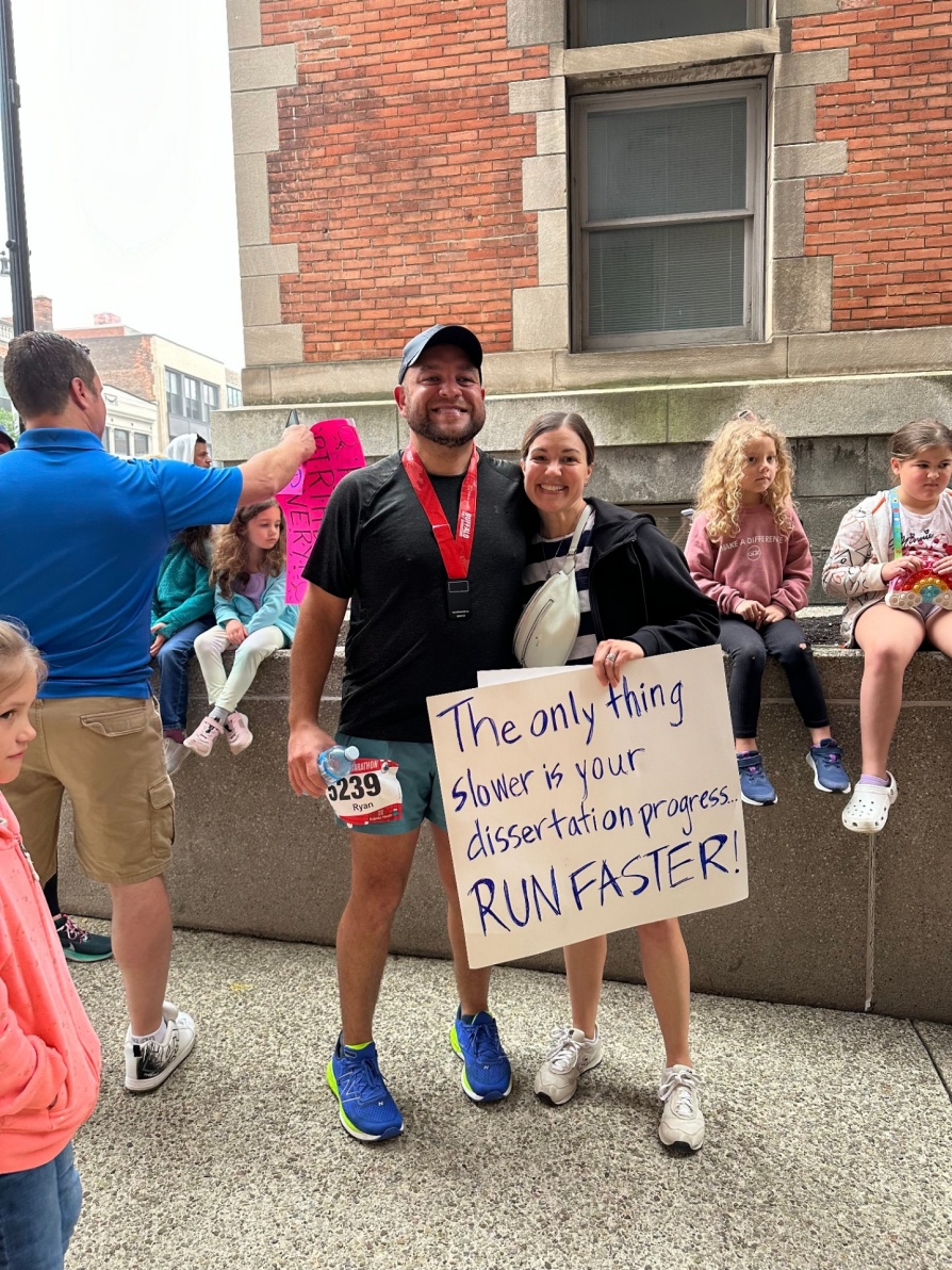 Zoom image: Ryan Taughrin and Danielle Lewis at a race. Danielle holding a sign that says "The only thing slower is your dissertation progress... Run faster!