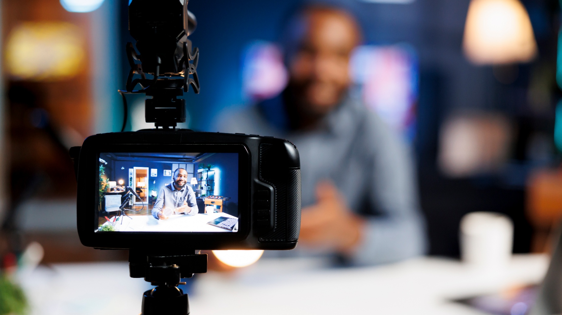 Video camera on a tripod recording a person seated at a desk, shown on the camera&rsquo;s display, with studio lighting in the background. 