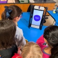Four elementary students gather around the M2 device as it begins a reflection session. The device displays and reads aloud: &ldquo;Okay, I am going to ask you some reflection questions. Respond out loud.&rdquo;. 