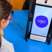 Four elementary students gather around the M2 device as it begins a reflection session. The device displays and reads aloud: &ldquo;Okay, I am going to ask you some reflection questions. Respond out loud.&rdquo;. 