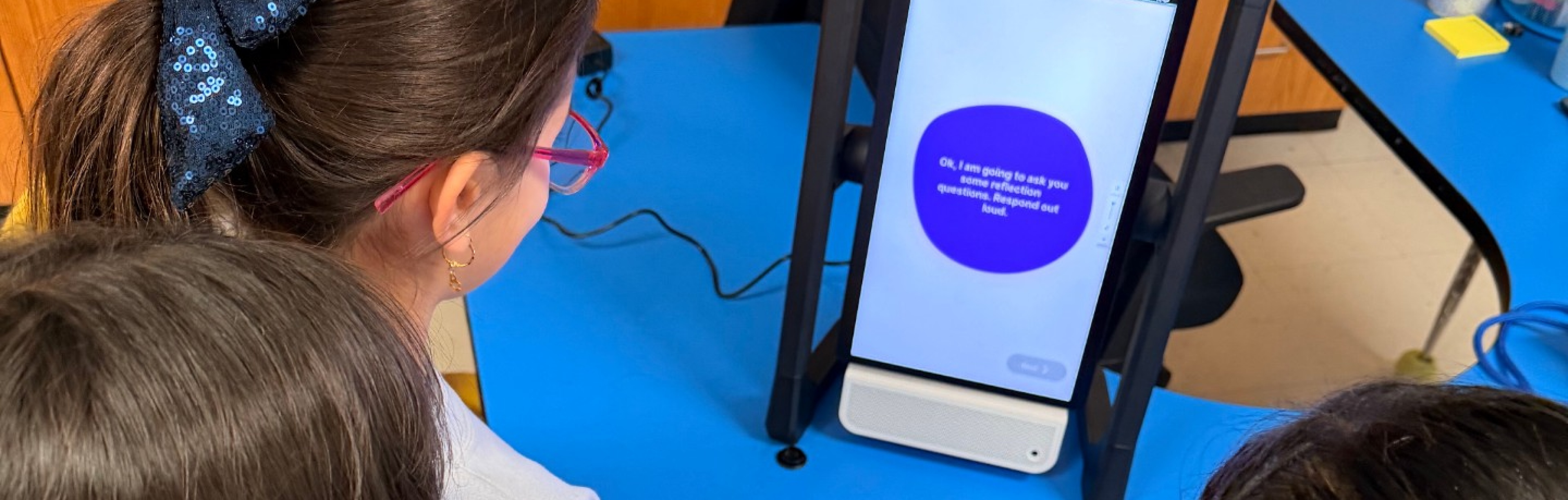 Four elementary students gather around the M2 device as it begins a reflection session. The device displays and reads aloud: &ldquo;Okay, I am going to ask you some reflection questions. Respond out loud.&rdquo;. 