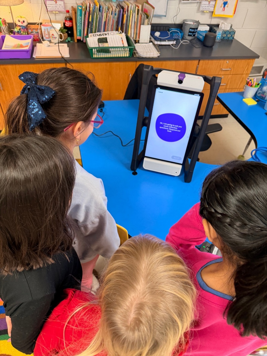 Four elementary students gather around the M2 device as it begins a reflection session. The device displays and reads aloud: &ldquo;Okay, I am going to ask you some reflection questions. Respond out loud.&rdquo;. 