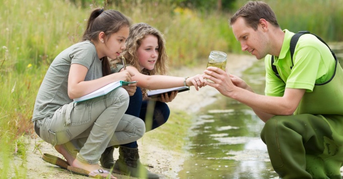 Teacher working in a creek teaching students science topics while showing a water sample.