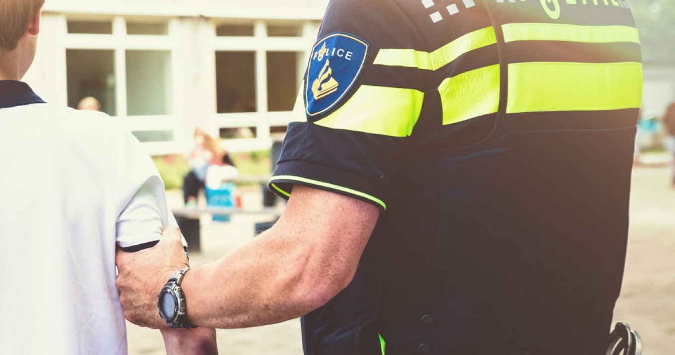 Policeman arresting young boy on schoolyard. Holding teenager at his arm. 