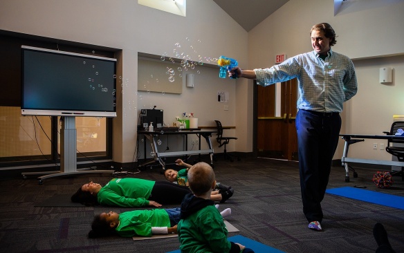 Zoom image: Yoga teacher filling a classroom with a K-2 class full of bubbles. 