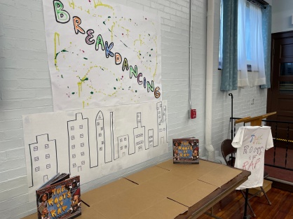 Zoom image: Student-created art and tee-shirts alongside &quot;The Roots of Rap&quot; book. 