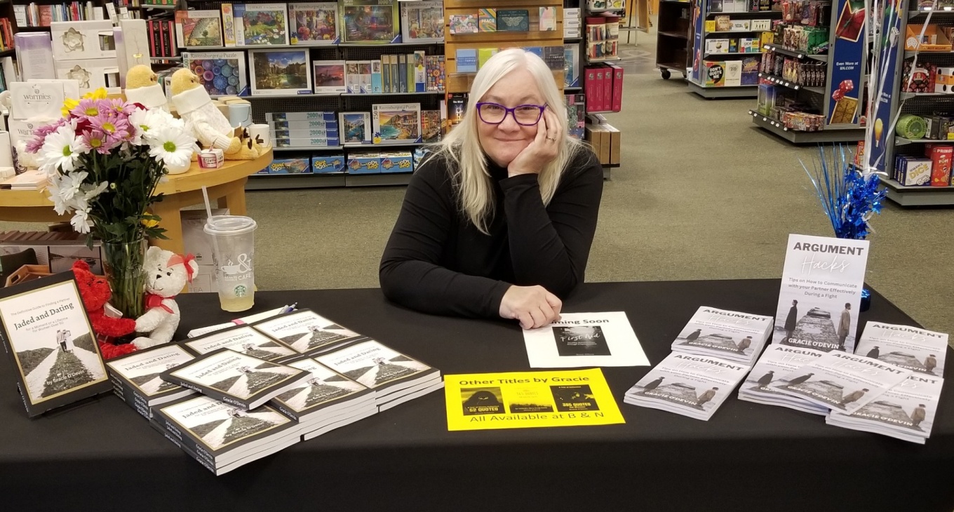 Kelly Devine poses at a signing table with her book “Jaded and Dating: The Definitive Guide to Finding a Partner for a Moment or a Lifetime for Women Over 50.". 