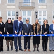 A group of seven people stand in a line outdoors in front of a large building, holding a long blue ribbon printed with the University at Buffalo logo. Each person holds ceremonial scissors as they participate in a ribbon‑cutting event. Snow is visible on the ground in the background near the building&rsquo;s entrance. 