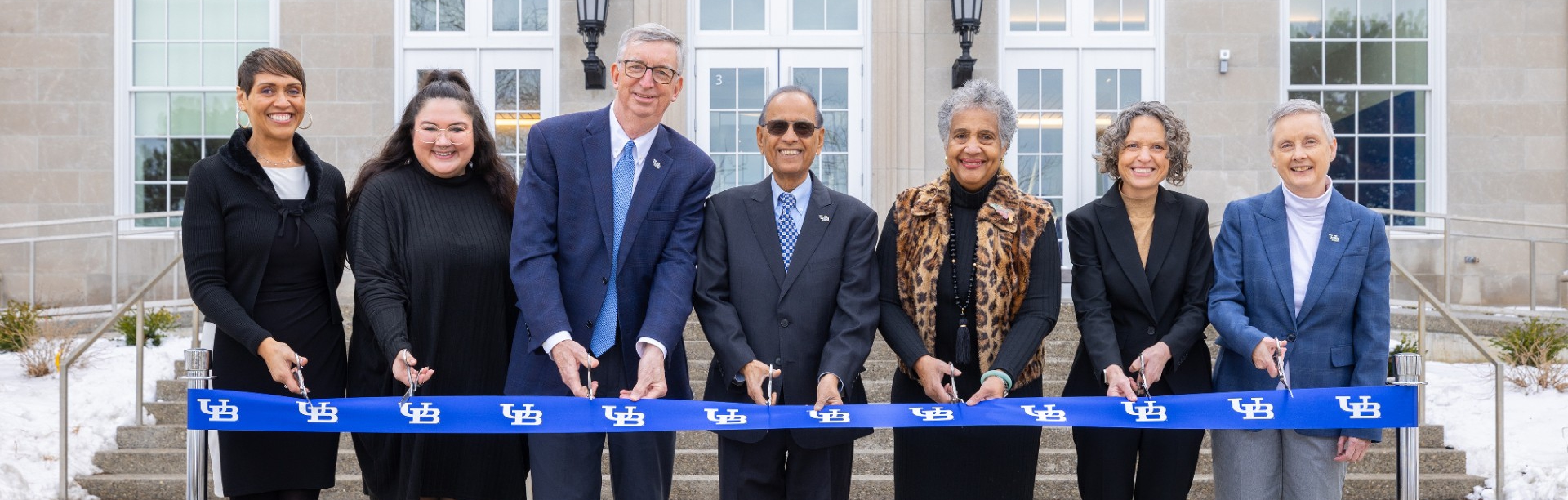 A group of seven people stand in a line outdoors in front of a large building, holding a long blue ribbon printed with the University at Buffalo logo. Each person holds ceremonial scissors as they participate in a ribbon‑cutting event. Snow is visible on the ground in the background near the building&rsquo;s entrance. 