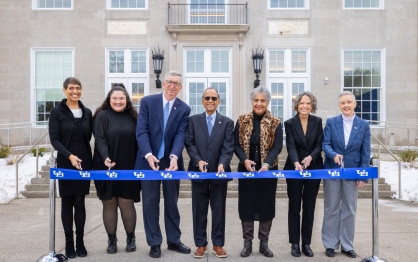 A group of seven people stand in a line outdoors in front of a large building, holding a long blue ribbon printed with the University at Buffalo logo. Each person holds ceremonial scissors as they participate in a ribbon‑cutting event. Snow is visible on the ground in the background near the building&rsquo;s entrance. 