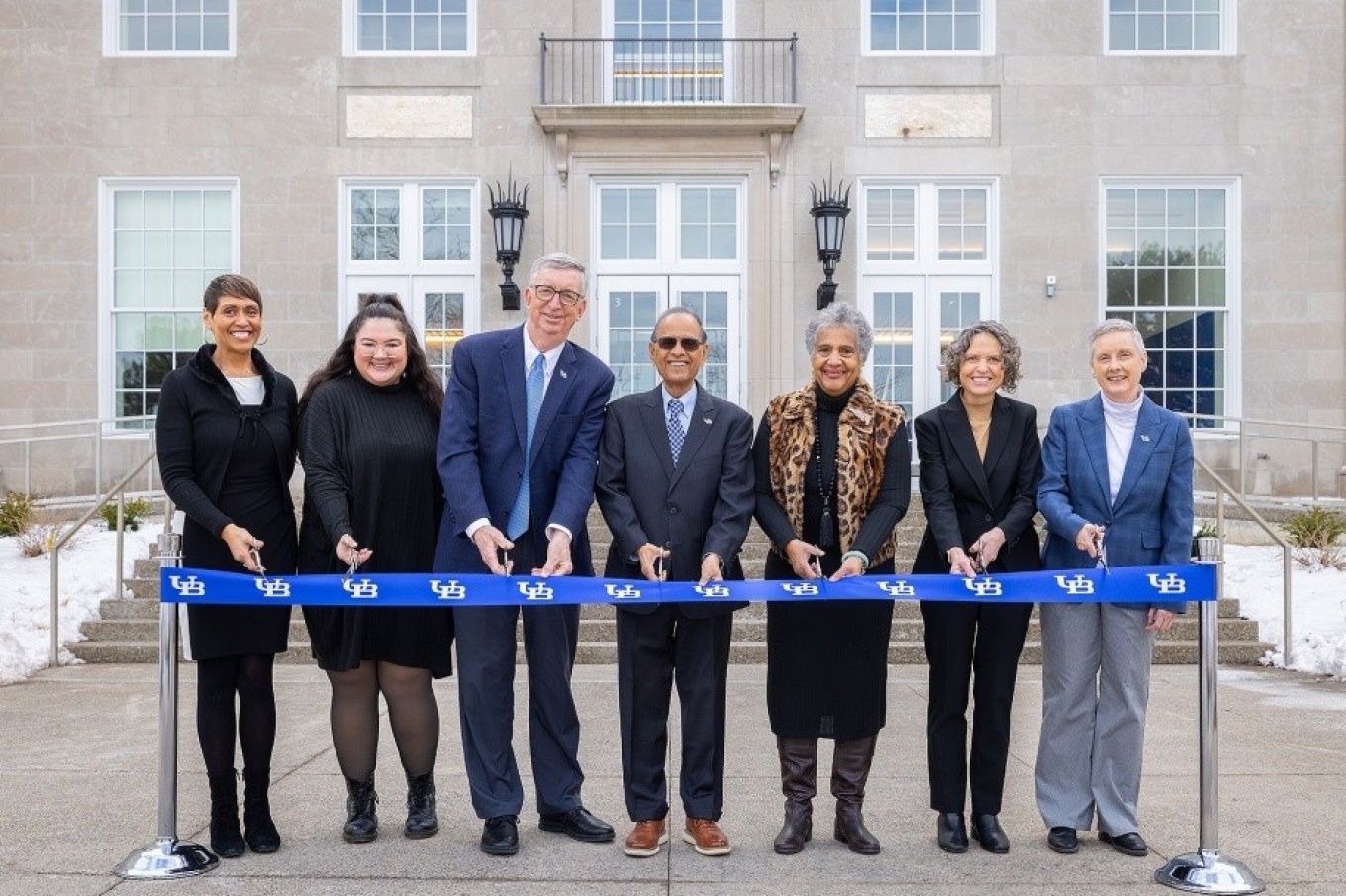 A group of seven people stand in a line outdoors in front of a large building, holding a long blue ribbon printed with the University at Buffalo logo. Each person holds ceremonial scissors as they participate in a ribbon‑cutting event. Snow is visible on the ground in the background near the building&rsquo;s entrance. 