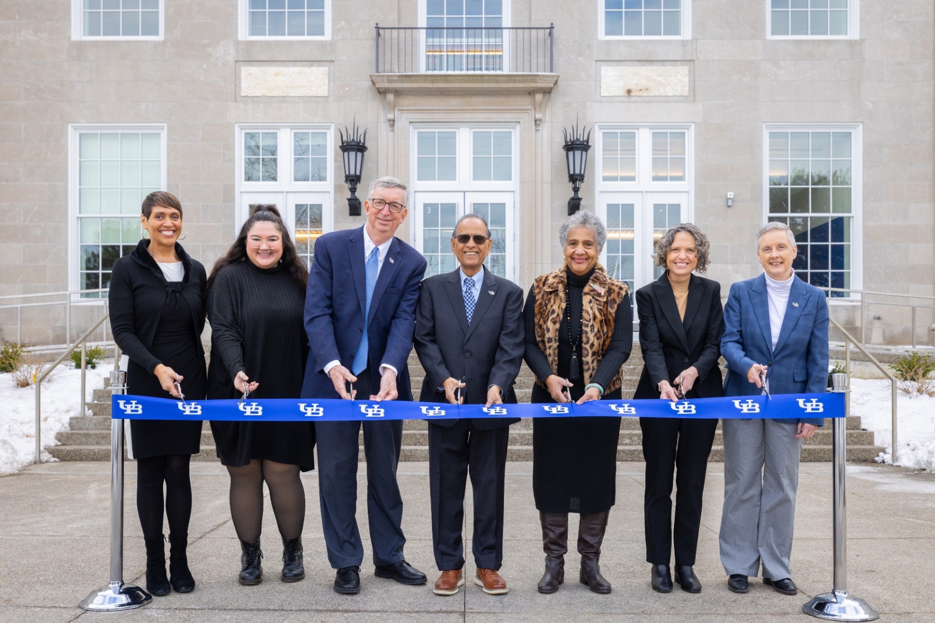 A group of seven people stand in a line outdoors in front of a large building, holding a long blue ribbon printed with the University at Buffalo logo. Each person holds ceremonial scissors as they participate in a ribbon‑cutting event. Snow is visible on the ground in the background near the building’s entrance.