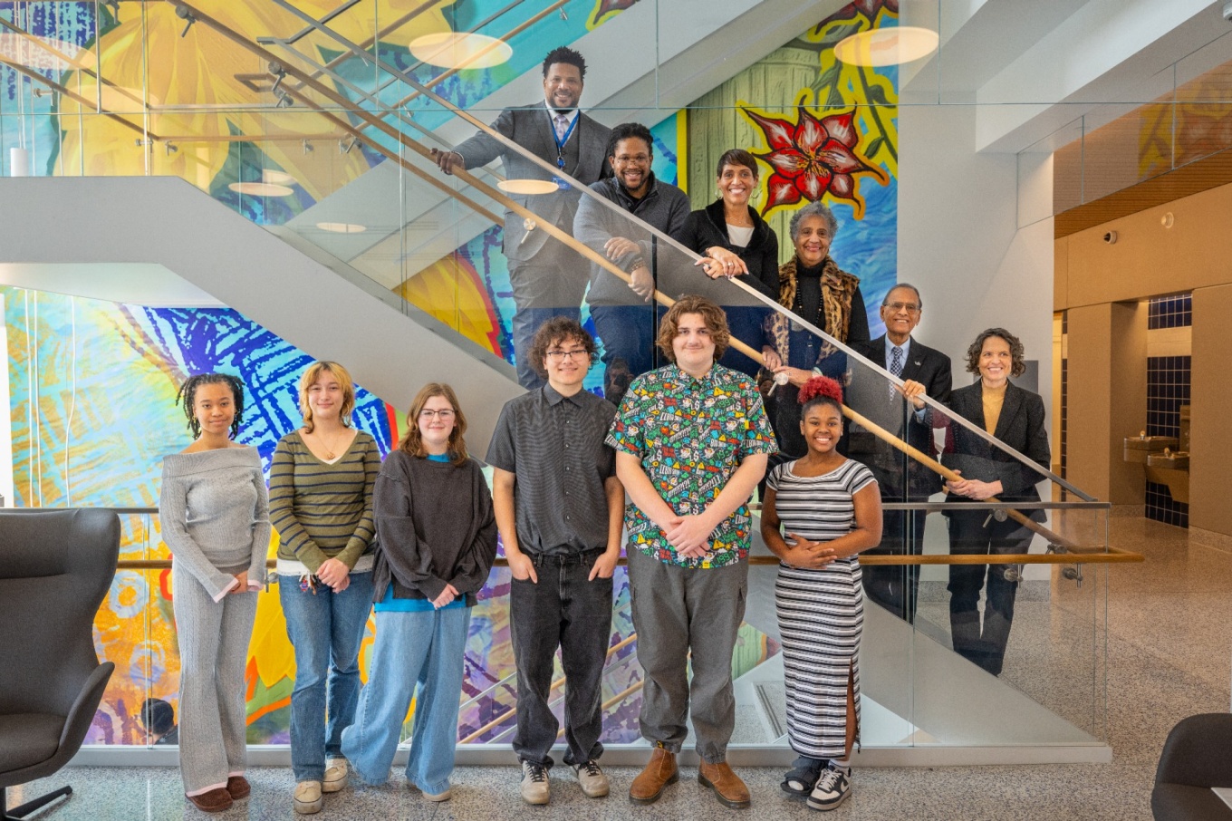 A group of twelve people pose on and in front of a modern indoor staircase. Some individuals stand on the steps behind a glass railing, while others stand on the floor level in front of the staircase. The background features large, brightly colored wall murals with abstract shapes and vivid patterns in blue, yellow, and red tones. The setting appears to be a contemporary building lobby or atrium with polished floors and modern lighting. 