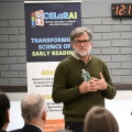 A person stands at the front of a room holding a water bottle and speaking. A banner to the left reads &ldquo;CELaRAI &ndash; Center for Early Literacy and Responsible AI&rdquo; with text about early reading research. To the right, a presentation slide is projected, showing the &ldquo;Digital Promise&rdquo; logo. An audience listens in the foreground. 