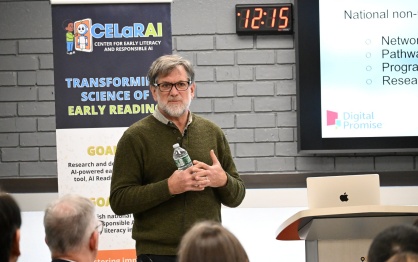 A person stands at the front of a room holding a water bottle and speaking. A banner to the left reads &ldquo;CELaRAI &ndash; Center for Early Literacy and Responsible AI&rdquo; with text about early reading research. To the right, a presentation slide is projected, showing the &ldquo;Digital Promise&rdquo; logo. An audience listens in the foreground. 
