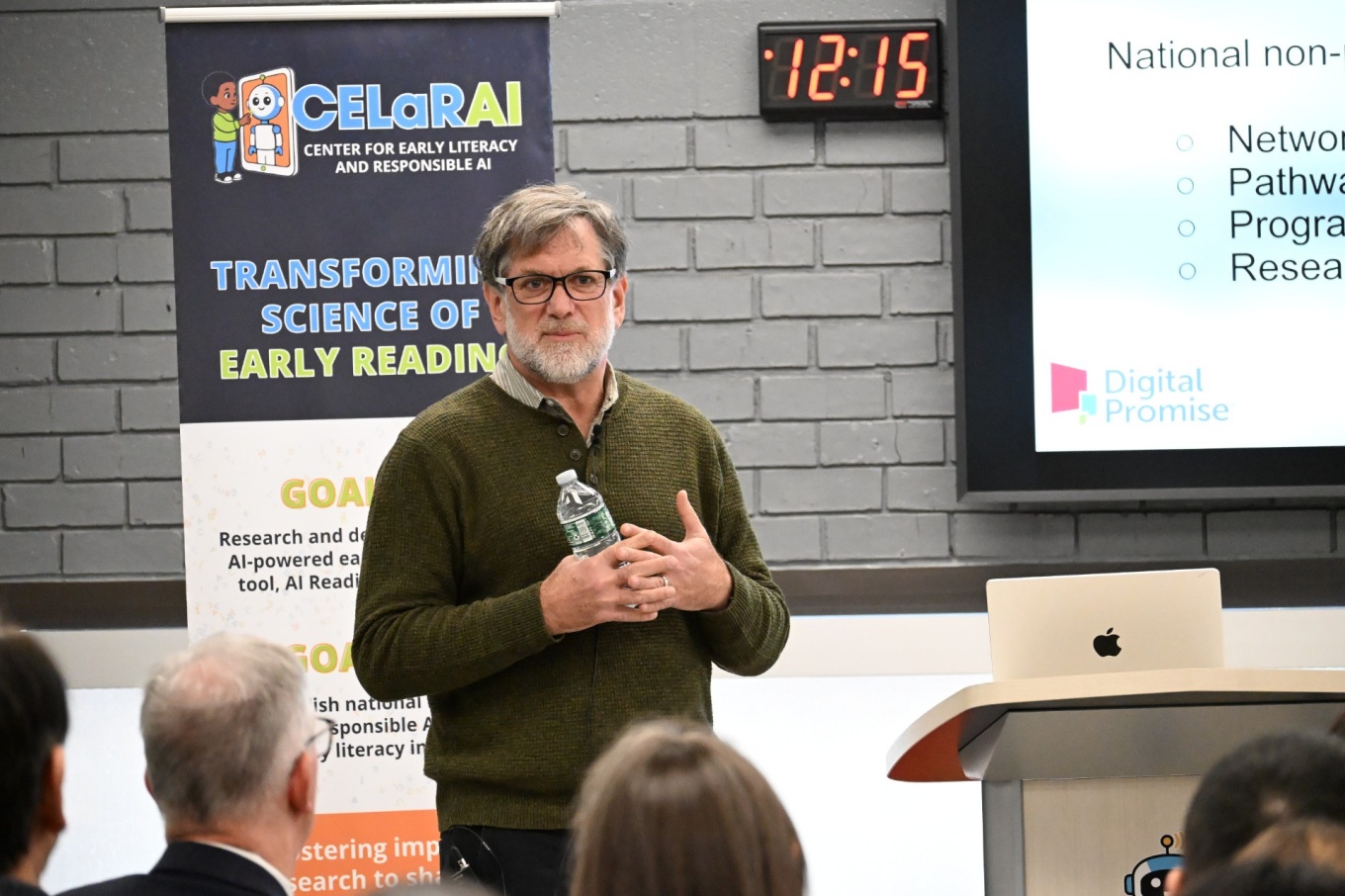 A person stands at the front of a room holding a water bottle and speaking. A banner to the left reads &ldquo;CELaRAI &ndash; Center for Early Literacy and Responsible AI&rdquo; with text about early reading research. To the right, a presentation slide is projected, showing the &ldquo;Digital Promise&rdquo; logo. An audience listens in the foreground. 