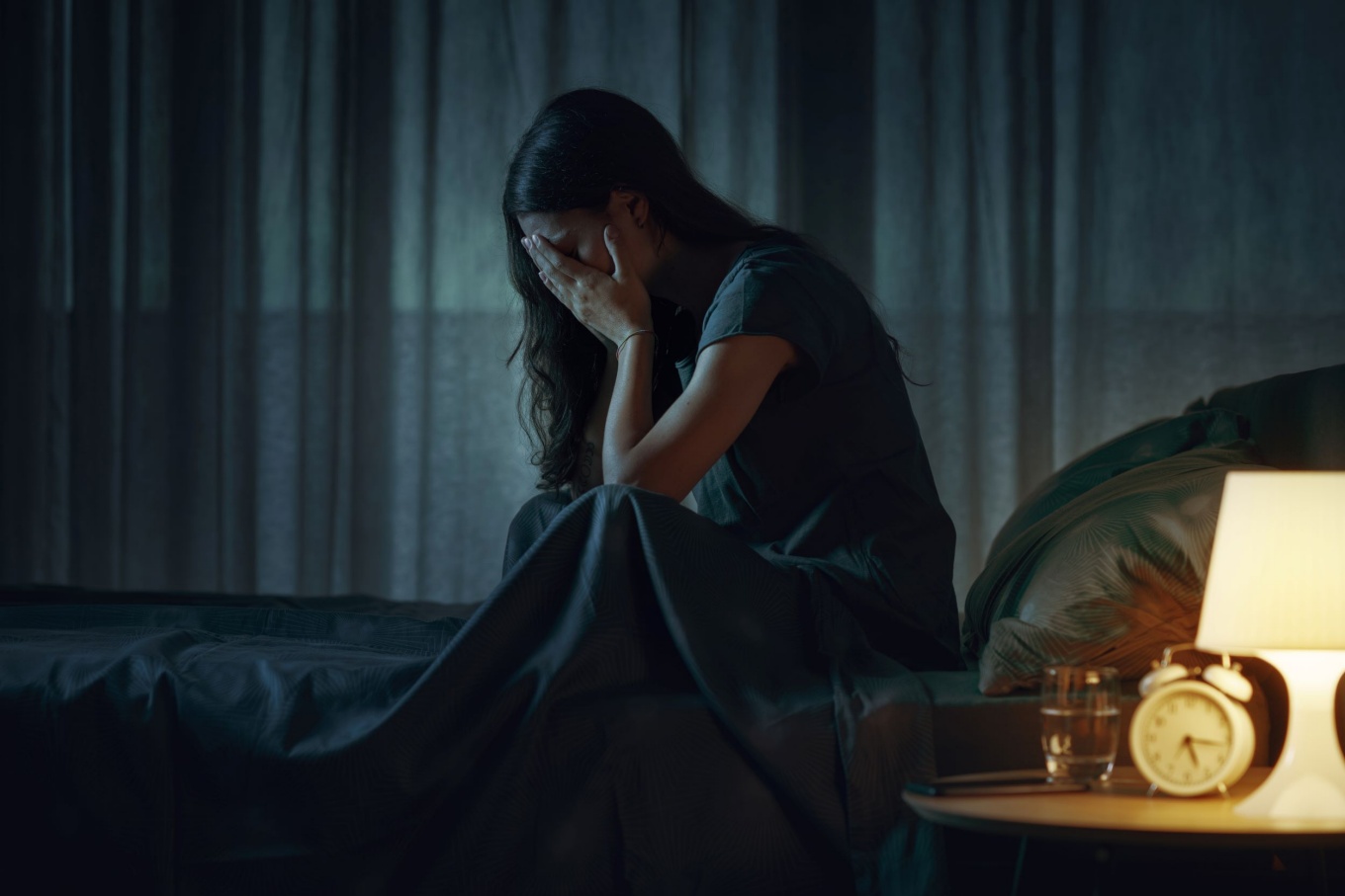 Person in dark room sitting on bed with face in hands. 