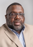 Portrait of a Black man with short hair and a beard wearing glasses, a blue shirt and light suit jacket. 