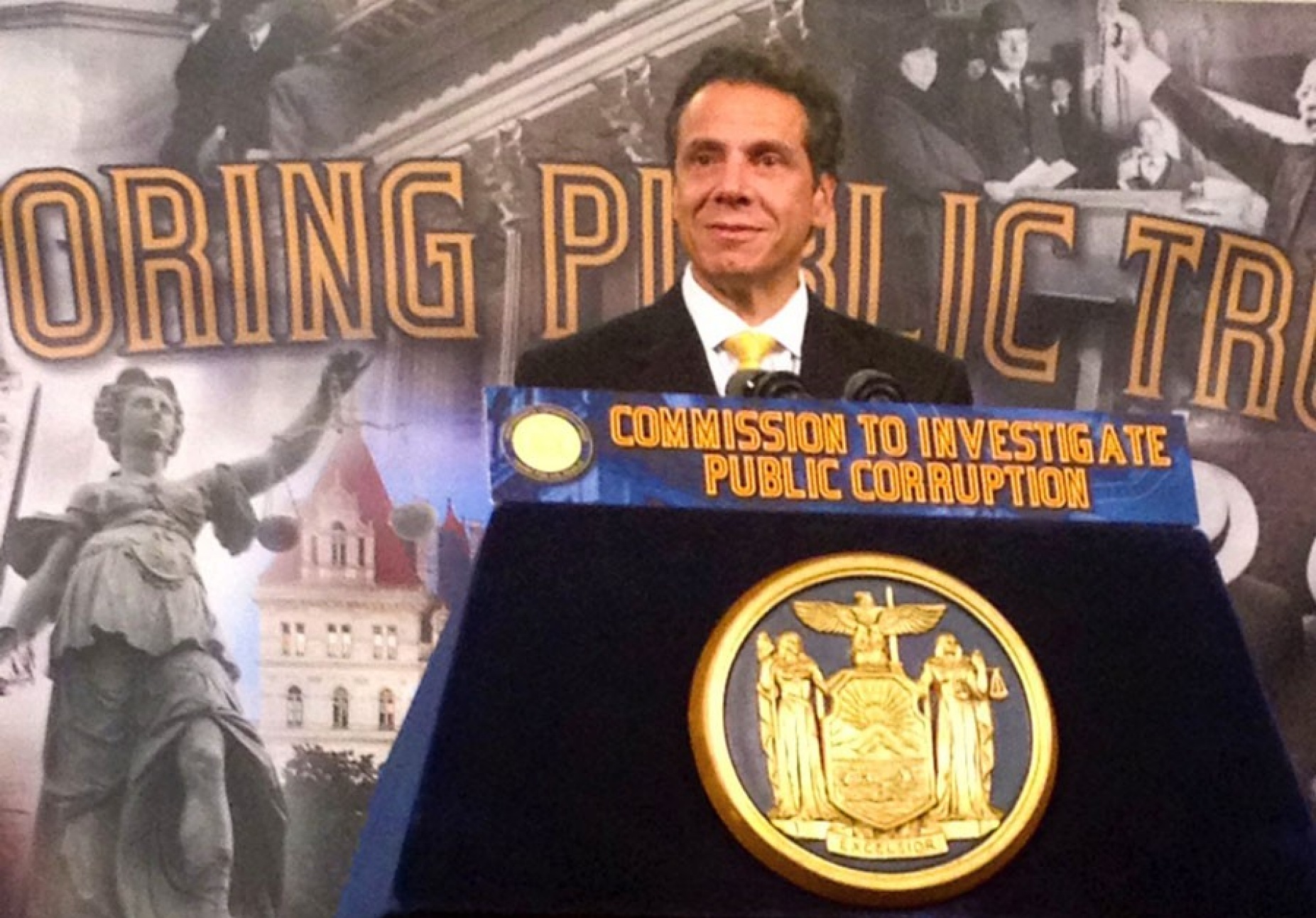Gov. Cuomo speaking from a podium. Signage on the podium reads, "Commission to investigate public corruption.". 