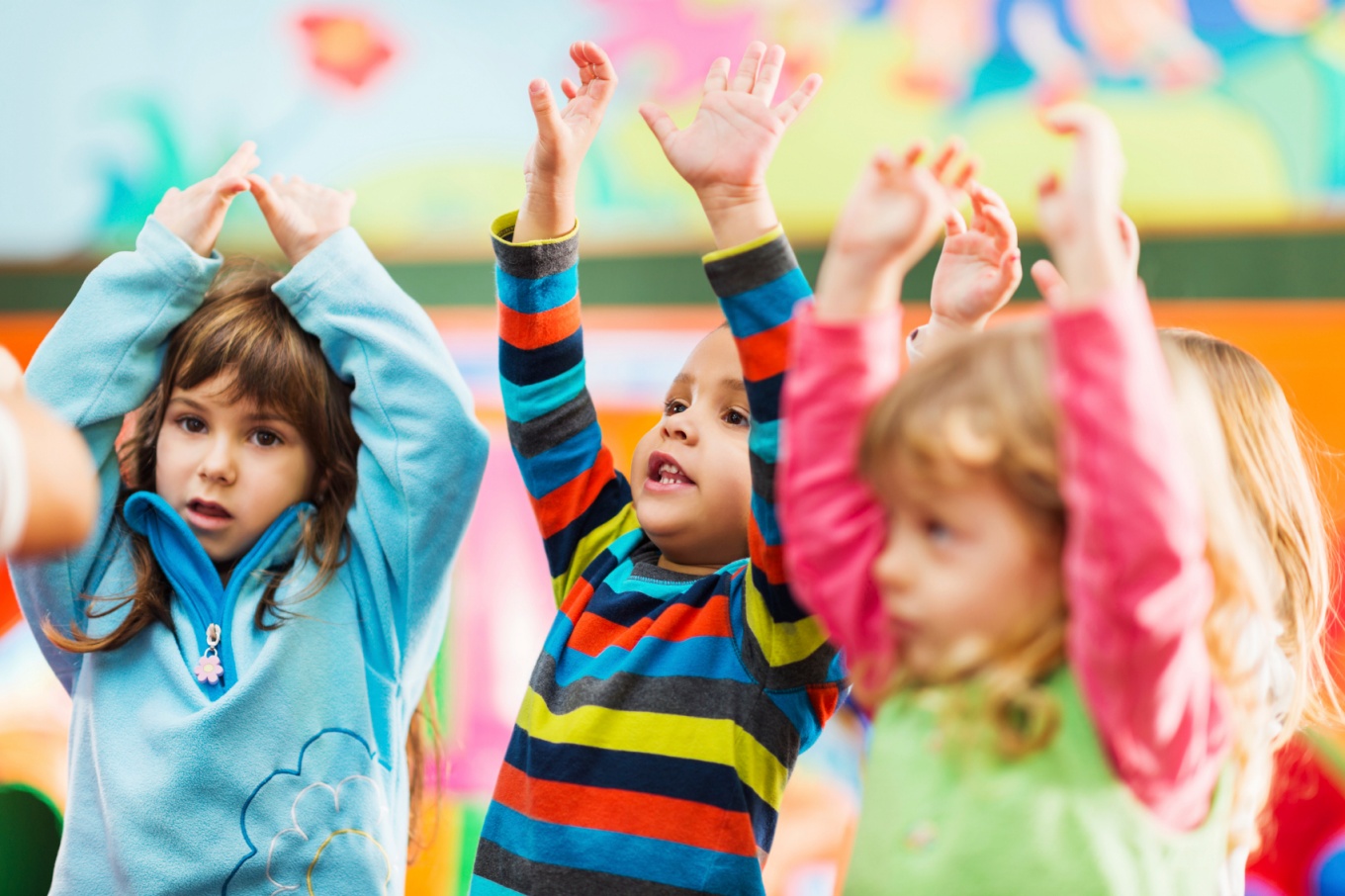 Three young children raising their hands in a classroom. 