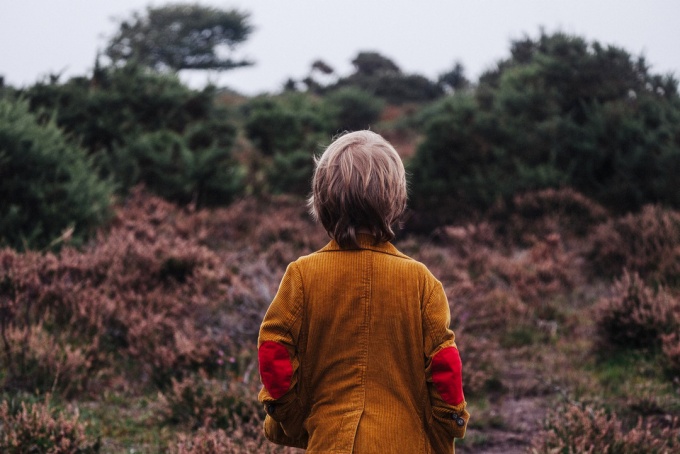 A boy wearing a tan jacket with red elbow patches stands with is back to the camera. 