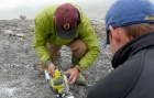 Two young men slicing a cylindrical sample of sediment encased in plastic. 