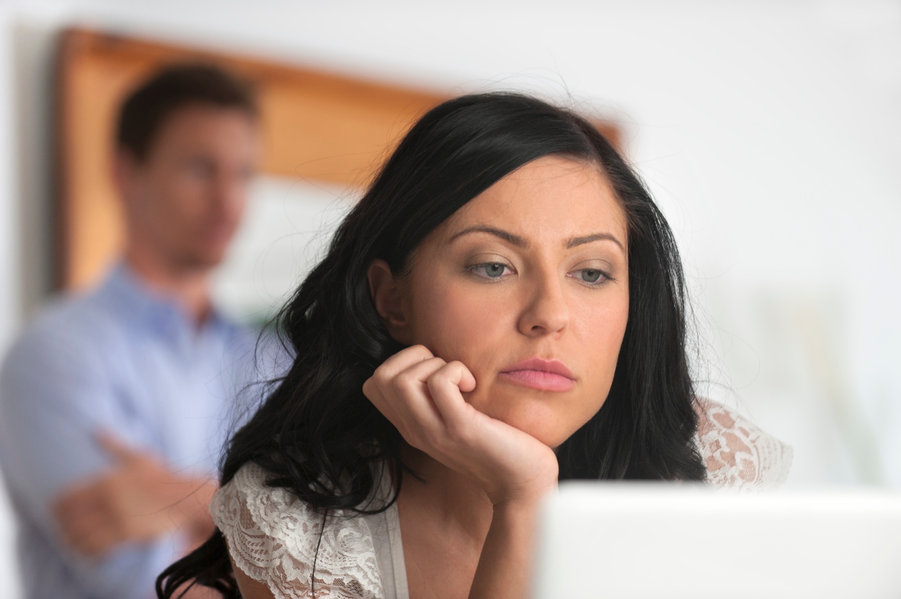 A woman stares at a laptop screen with an out of focus man standing in the background. 