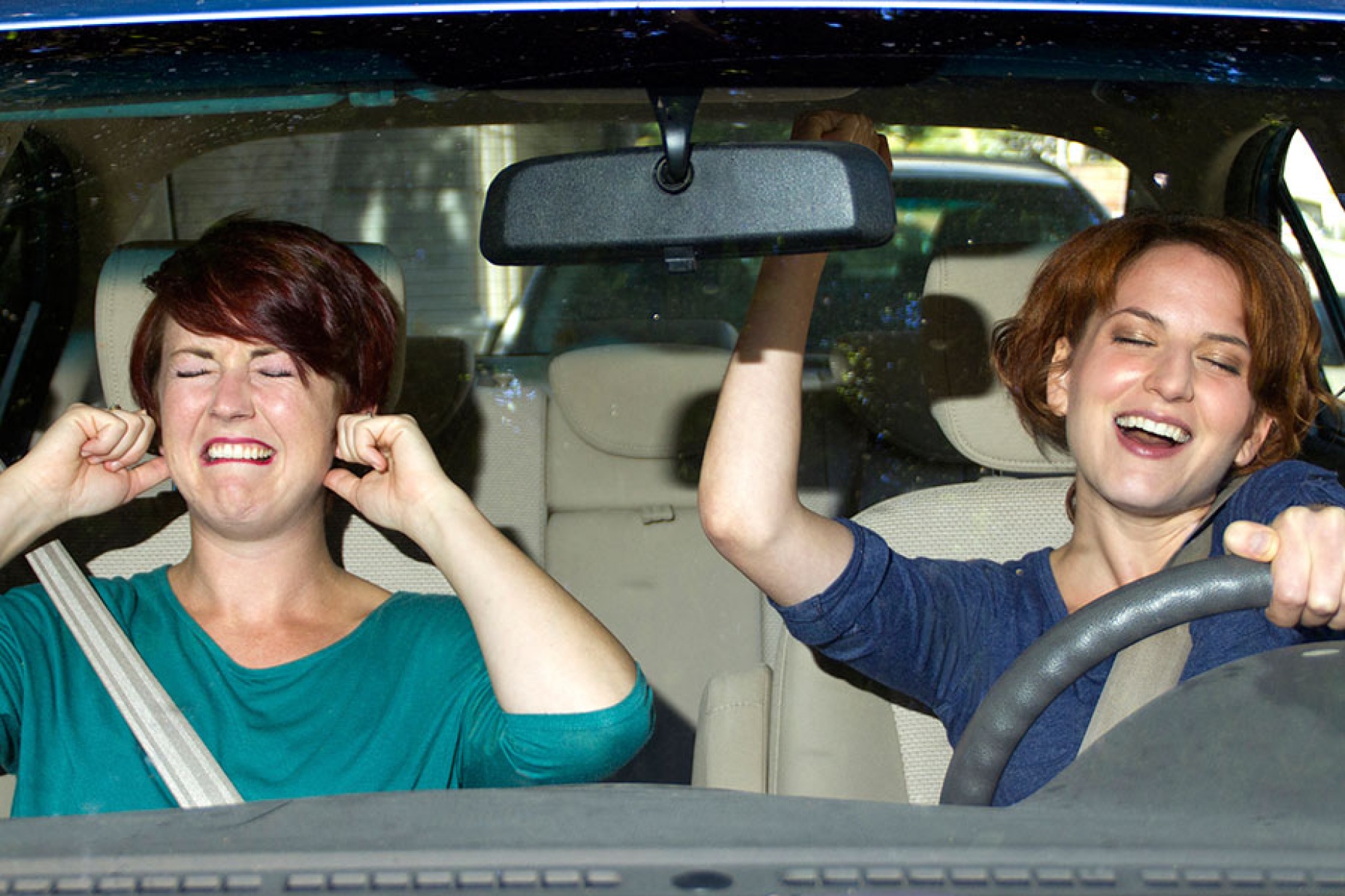 A woman sings badly in the car as her passenger hold her ears and grimaces.