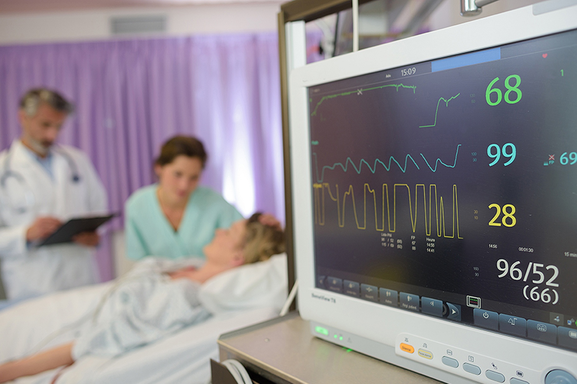 Scene of a hospital room with a hospital monitor featured prominently in the foreground and medical staff attending to a patient in the background. 