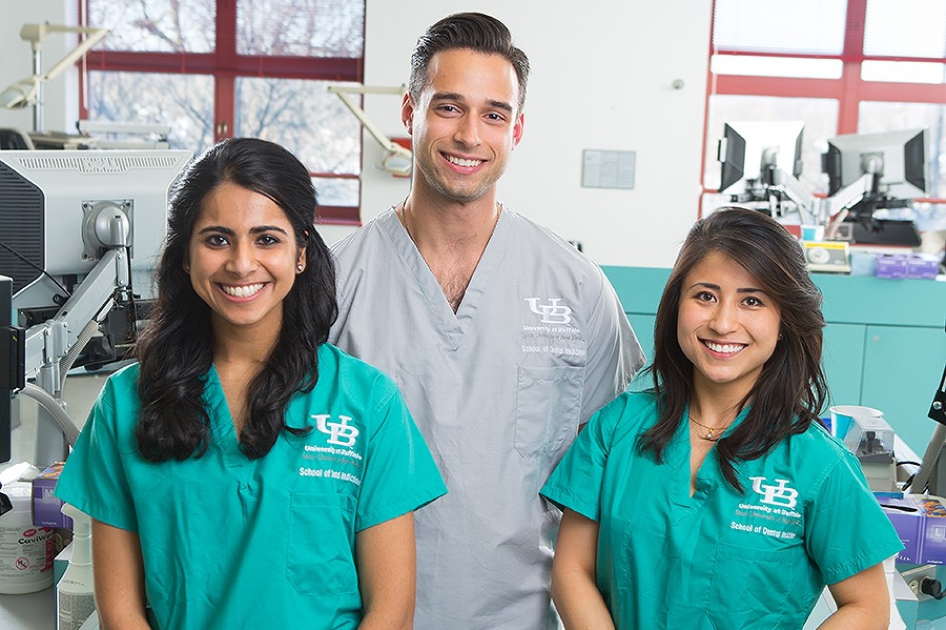 Three students in grey or green scrubs smiling in dental clinic.