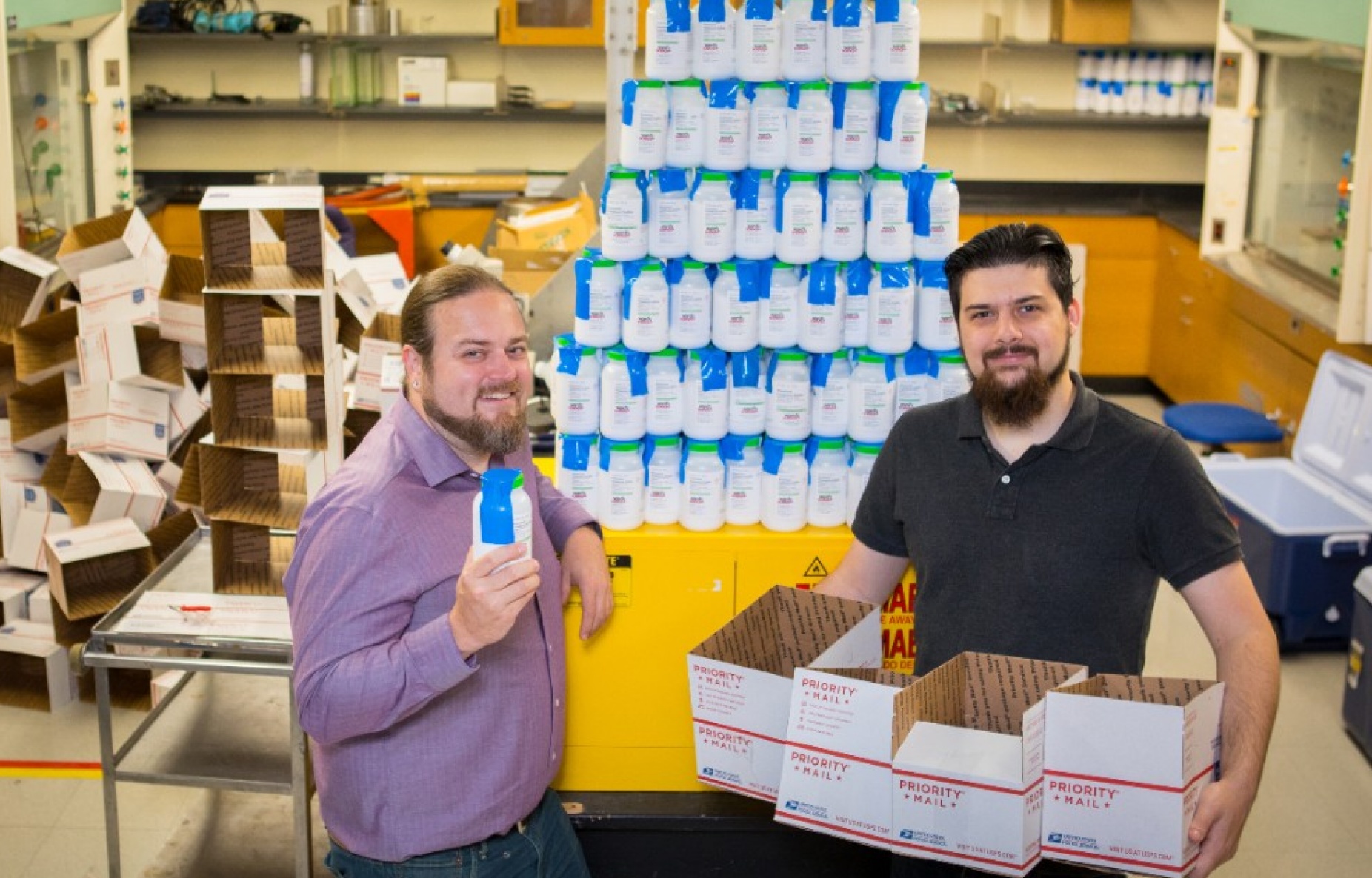 Two people standing in front of a tall triangular stack of bottles holding a crystal-growing chemical. 