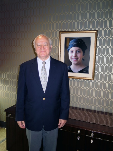 Allen Barnett wearing a suit and standing in front of a portrait of his granddaughter, Carly. 