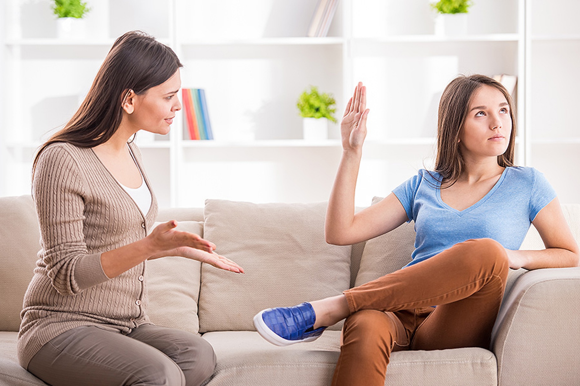 Teen girl is showing stop gesture to angry mother while sitting on sofa at home. 