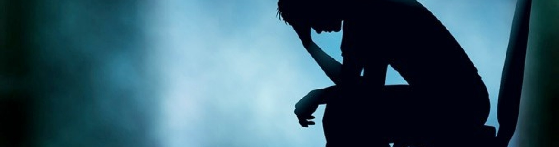 A silhouette of a person sitting on a chair with their head in the hands and fog in the background. 