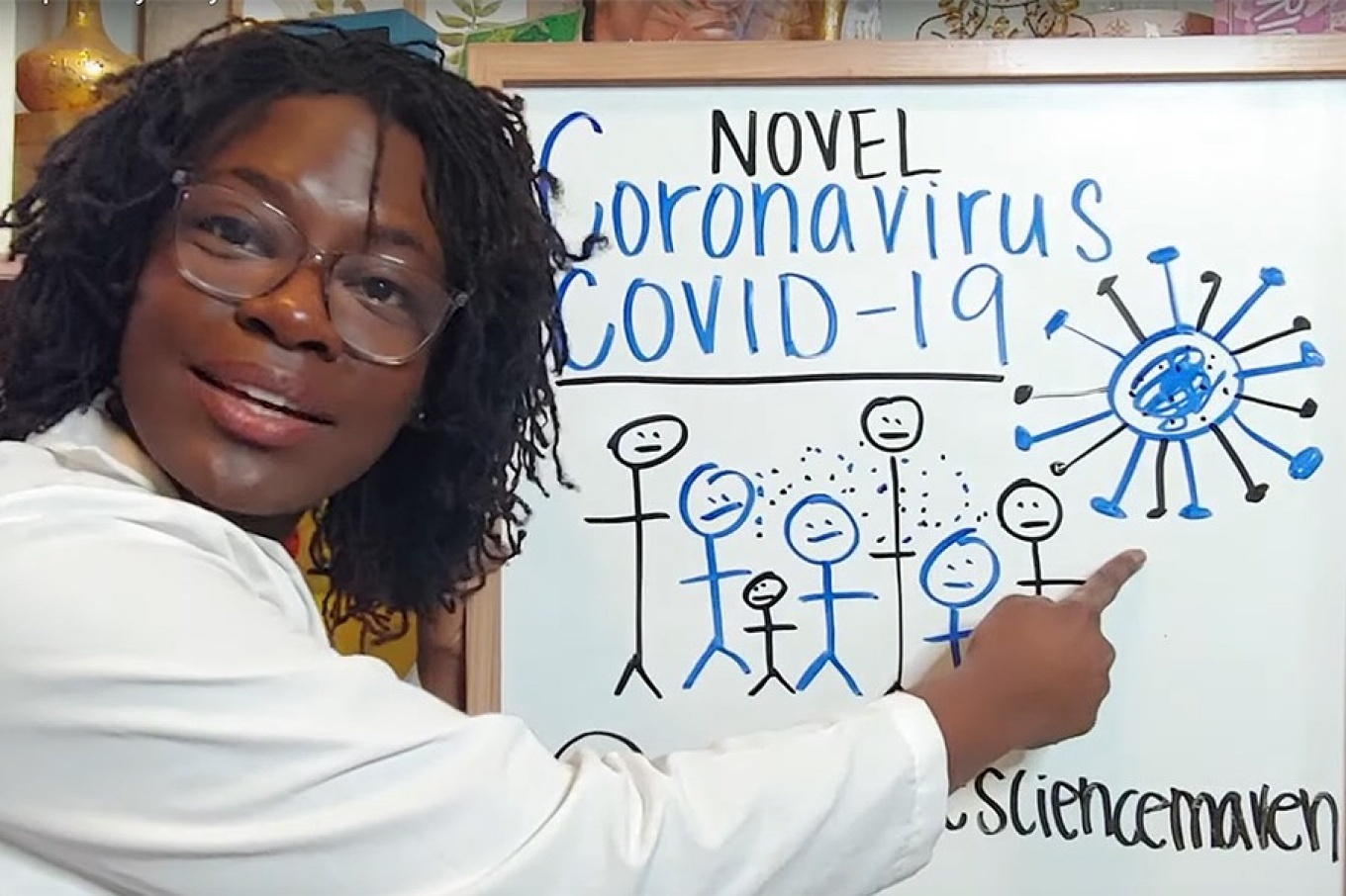 Raven Baxter in white lab coat points to whiteboard with details on coronavirus. 