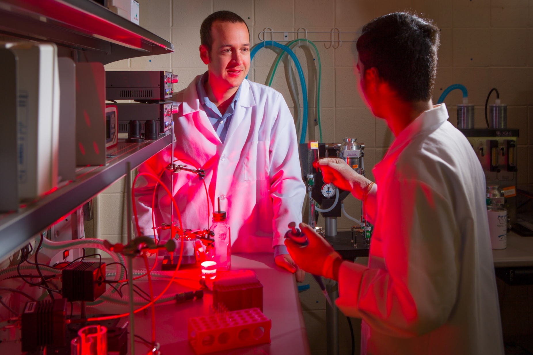 UB researcher Jon Lovell stands in his lab in a white coat talking to a student researcher while a red light from a machine colors the room. 