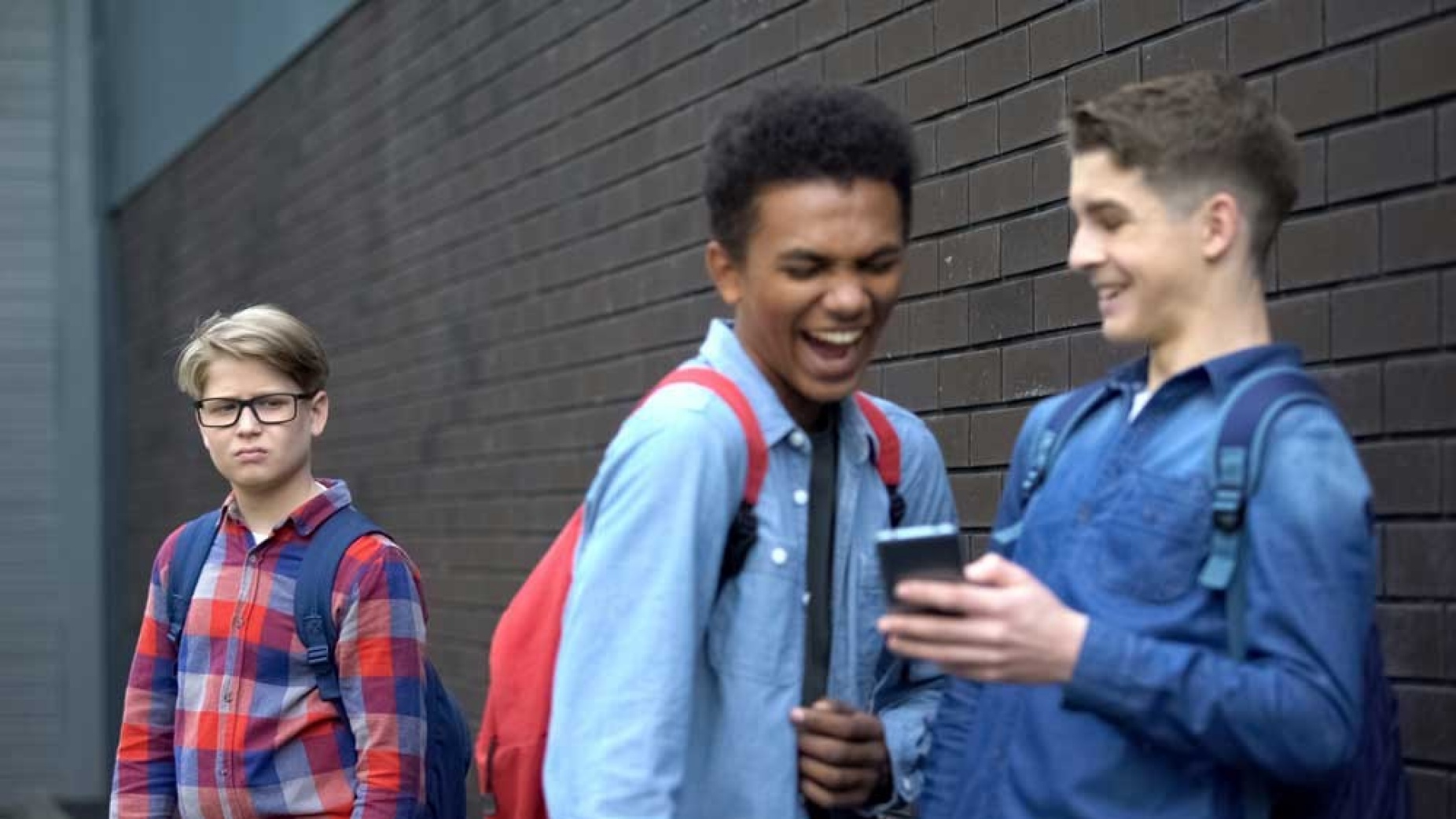 Two school-age boys looking at a phone and laughing in front of another boy who is visibly upset. 