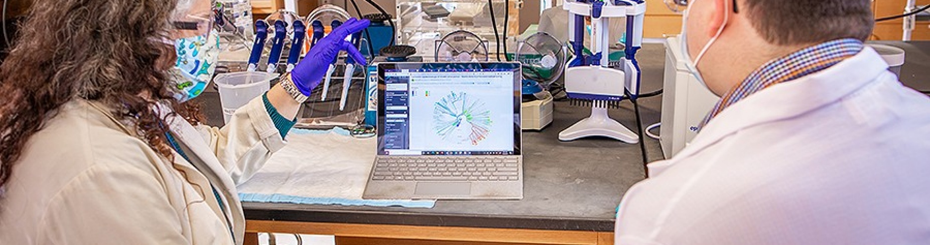 Jennifer Surtees and Donald Yergeau, in white coats and seated, with their backs to camera, examine data on computer screen.
